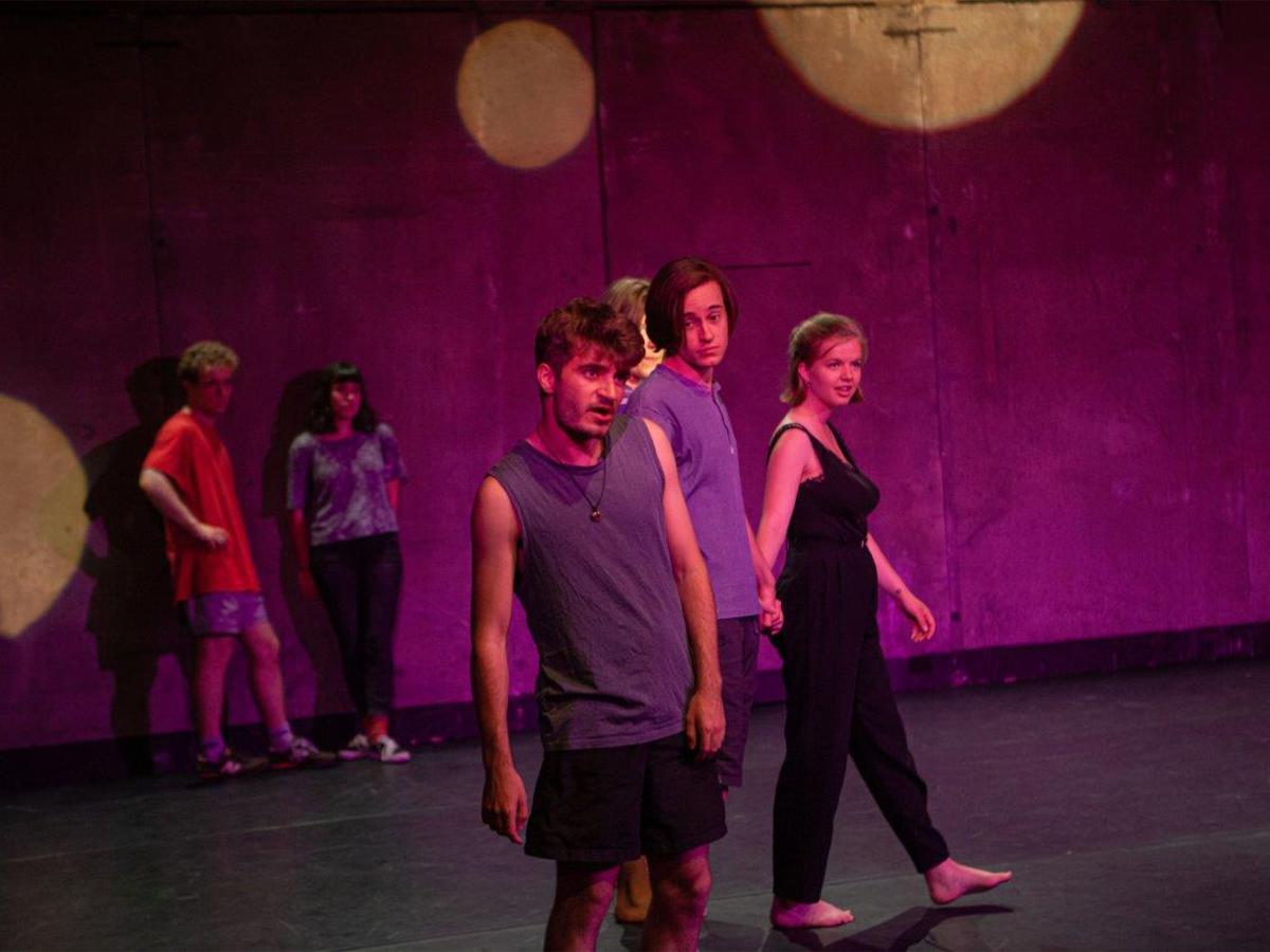Mehrere junge Menschen stehen und bewegen sich auf einer Bühne im pink-violetten Scheinwerferlicht, als Teil einer Theater- oder Tanzprobe.