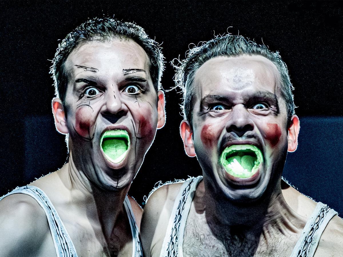 Nils Kahnwald und Kristof Van Boven nebeneinander in Nahaufnahme. Sie tragen weiße Unterhemden mit vielen kleinen Löchern und schauen mit verzerrtem Gesicht in die Kamera. Ihre Wangen sind grob mit roter Farbe bemalt, die Nase und Augenbrauen von Nils Kahnwald sind mit schwarzem Stift grob skizziert. Ihre Münder sind weit aufgerissen, es leuchtet grellgrün aus ihnen.