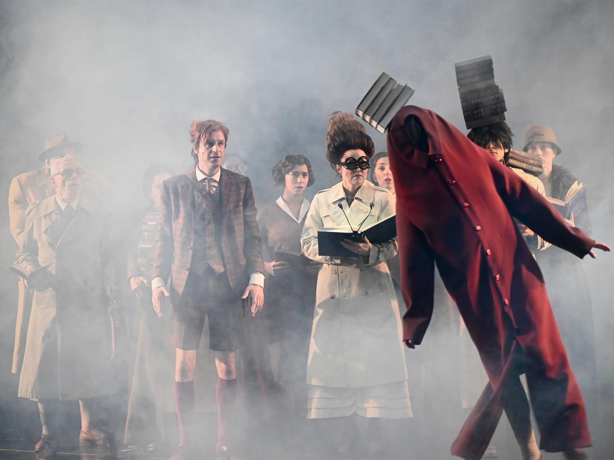 Bühnensituation: Das Ensemble steht als Gruppe zusammen in zwei Reihen. Sie tragen lange braune und beige Trenchcoats und halten Bücher in der Hand. Eine Person balanciert einen Stapel Bücher auf dem Kopf und der Schulter. Sie schauen auf eine Figur im Vordergrund. Sie trägt ein körperlanges rotes zugeknöpftes Hemd mit langen Ärmeln, das weit oberhalb des Kopfes endet, sodass kein Kopf zu sehen ist. Die Figur steht schräg, ebenfalls mit einem schiefen Stapel Bücher auf dem Kopf. Die Szenerie ist in dichten